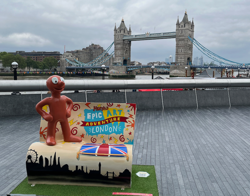 Morph Over, There's Room For Two, a sculpture of Morph standing on a bench, with Tower Bridge in the background. The space next to Morph on the bench has the colourful logo for Morph's Big Art Adventure London on the back rest, and a Union Jack cushion painted on the seat. The bottom section of the bench has a silhouette of the London skyline going across it.