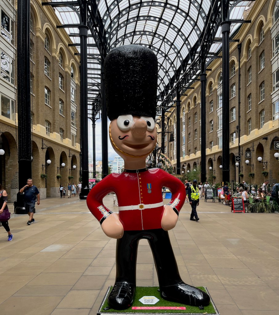 On Guard, a statue of Morph dressed as a royal guard, with a black furry busby hat held in place by a gold chinstrap, a red jacket and black trousers.