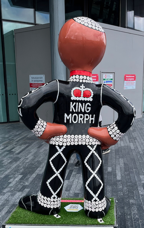The back of the Pearly King Morph statue, with white lettering on his black suit that says King Morph, below an image of a crown with a red centre surrounded by a border of white pearl buttons.