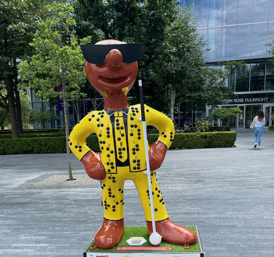 Power of Morphing Communication, a statue of Morph wearing a yellow suit and tie covered in black braille lettering, with printed letters to say what each one is. Morph is also wearing dark sunglasses and holding a long white cane.