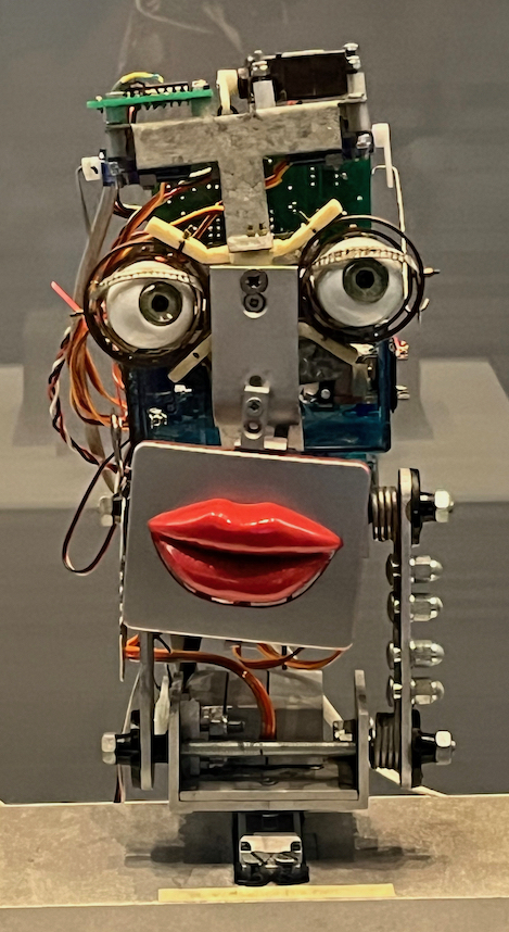 Inkha Reactive Robotic Head. This is a robotic head made up of a mish-mash of parts and circuits, including a metal panel with a pair of lips below 2 fake eyes.