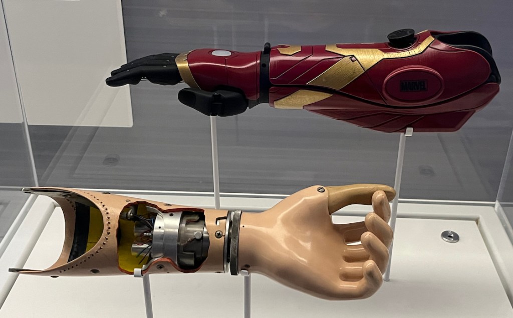 Two prosthetic arms, one red in colour with gold decorations, the other having the colour of human skin with a large hole in the wrist exposing the inner workings.