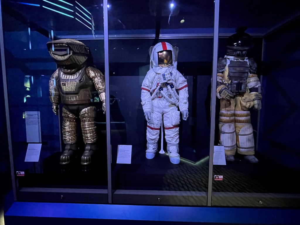 Three spacesuits, in gold and white colours. They are the gold spacesuit from a film called Sunshine, Gene Cernan’s Extravehicular Mobility Unit Suit, and a Nostromo Crew Spacesuit from the film Alien.