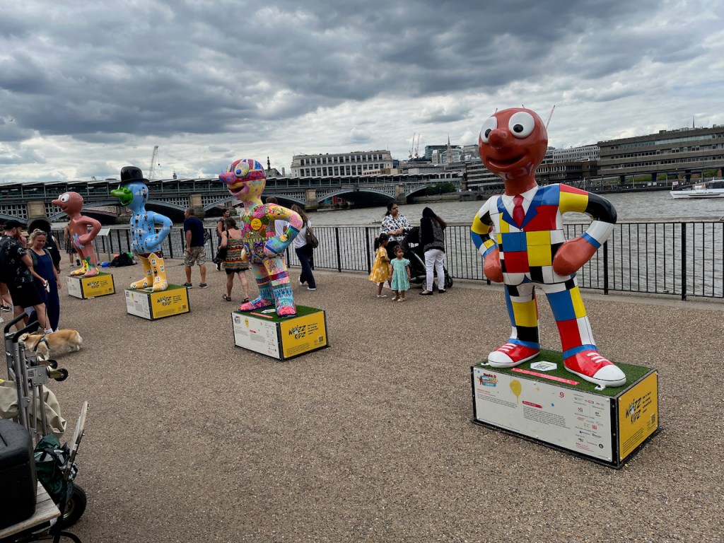 4 colourful sculptures of Morph lined up in a row on the South Bank of the River Thames, with Blackfriars Bridge in the background.