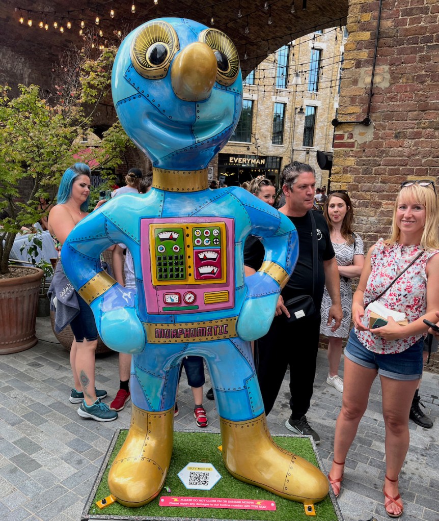 Totally Morphomatic, a statue of Morph looking like a robot, made up of panels in varying shades of blue bolted together. There are buttons and dials on his chest, and he has large gold eyes, a gold nose, gold bands around his neck and wrists, and gold shoes.