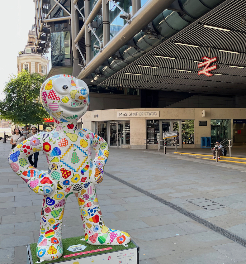 Apples and Pears, a white statue of Morph covered in drawings of the fruits in the title, all filled with a myriad of different colours and patterns. The entrance to Cannon Street station is to the right of the statue, next to a branch of M&S Simply Food in the background.