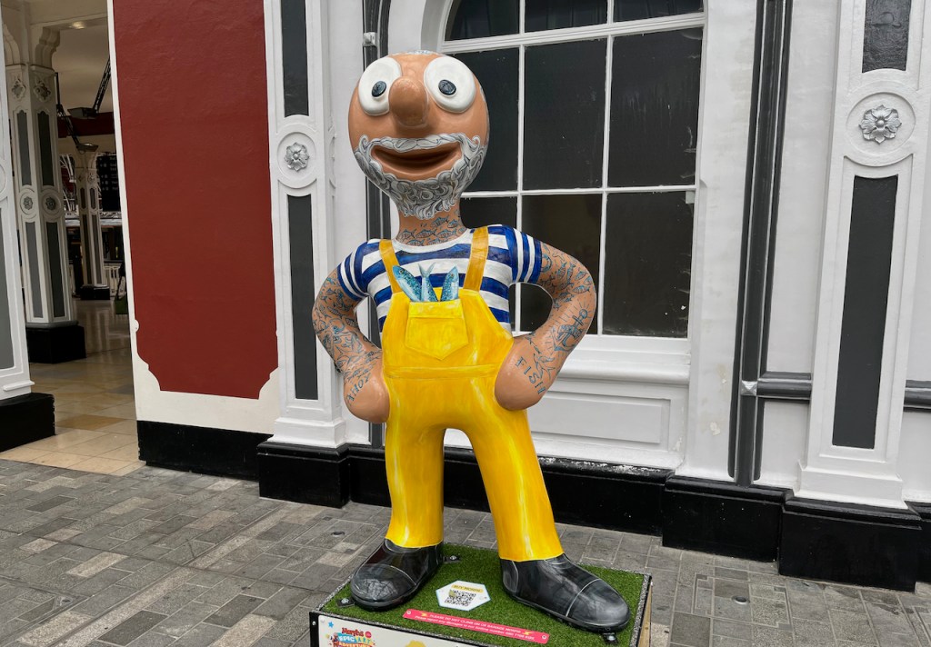 Fish Ahoy, a statue of Morph dressed as a fisherman, wearing a blue and white striped t-shirt, over which is a yellow outfit that straps over his shoulders, effectively combining an apron with trousers, with fish poking out of the central chest pocket. He also has a curly white beard and moustache, and small fish tattooed on his neck.