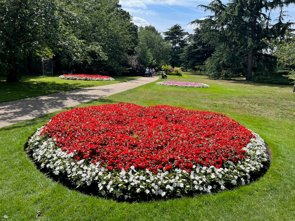 Large round flowerbed filled with red flowers, surrounded by a border of white flowers. Similar flowerbeds with pink or red flowers in the centre can be seen further along the grass on each side of the path in the distance.