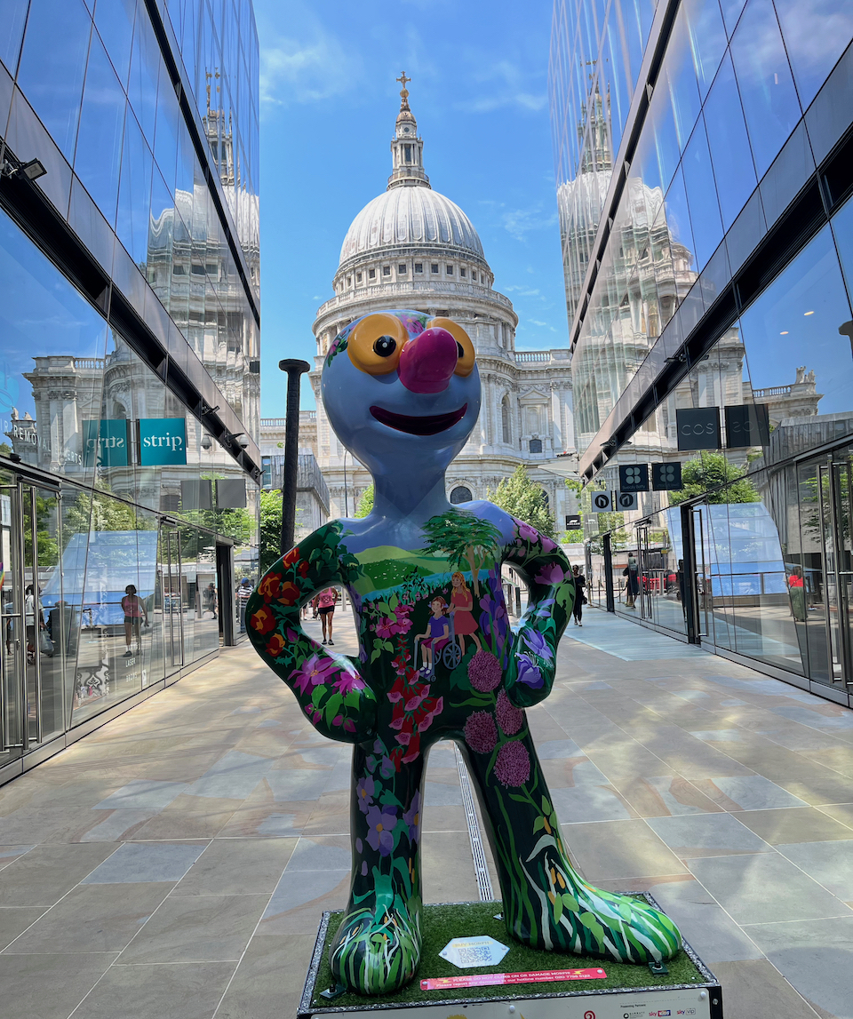 Freedom, a blue statue of Morph on which is a colourful painting of a lady pushing a woman in a wheelchair through a park full of colourful flowers, trees and greenery, with a river flowing past behind them. Behind the statue, St Paul's Cathedral stands tall in the background.