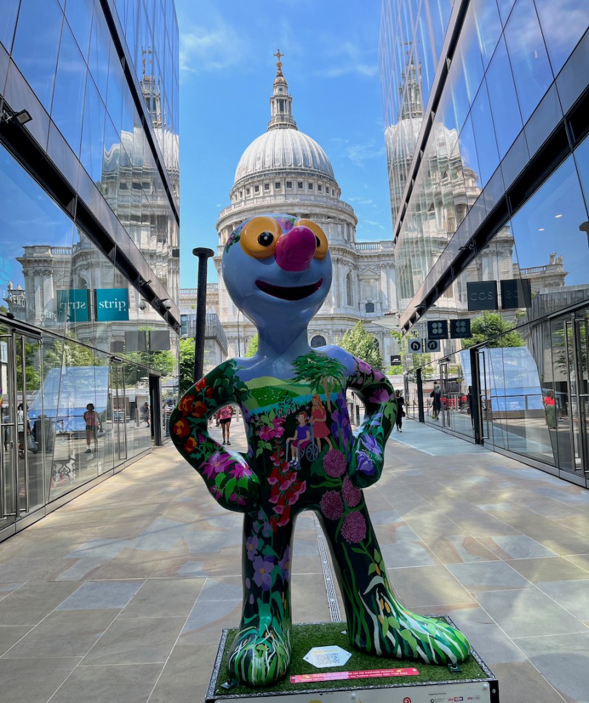 Freedom, a blue statue of Morph on which is a colourful painting of a lady pushing a woman in a wheelchair through a park full of colourful flowers, trees and greenery, with a river flowing past behind them. Behind the statue, St Paul's Cathedral stands tall in the background.