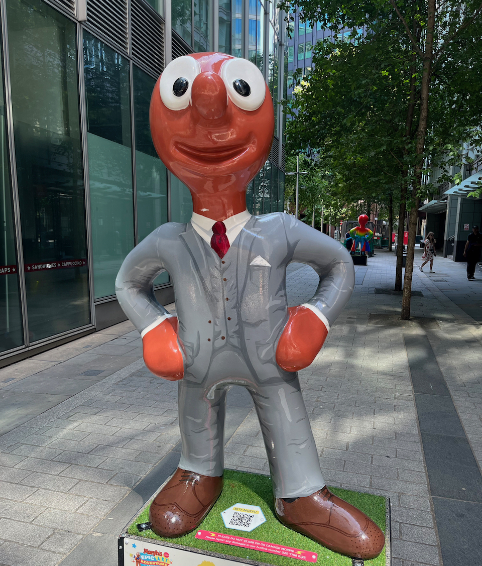 Gentle-morph, a statue of Morph dressed in a smart grey suit, over a white shirt with a red tie, and brown shoes.