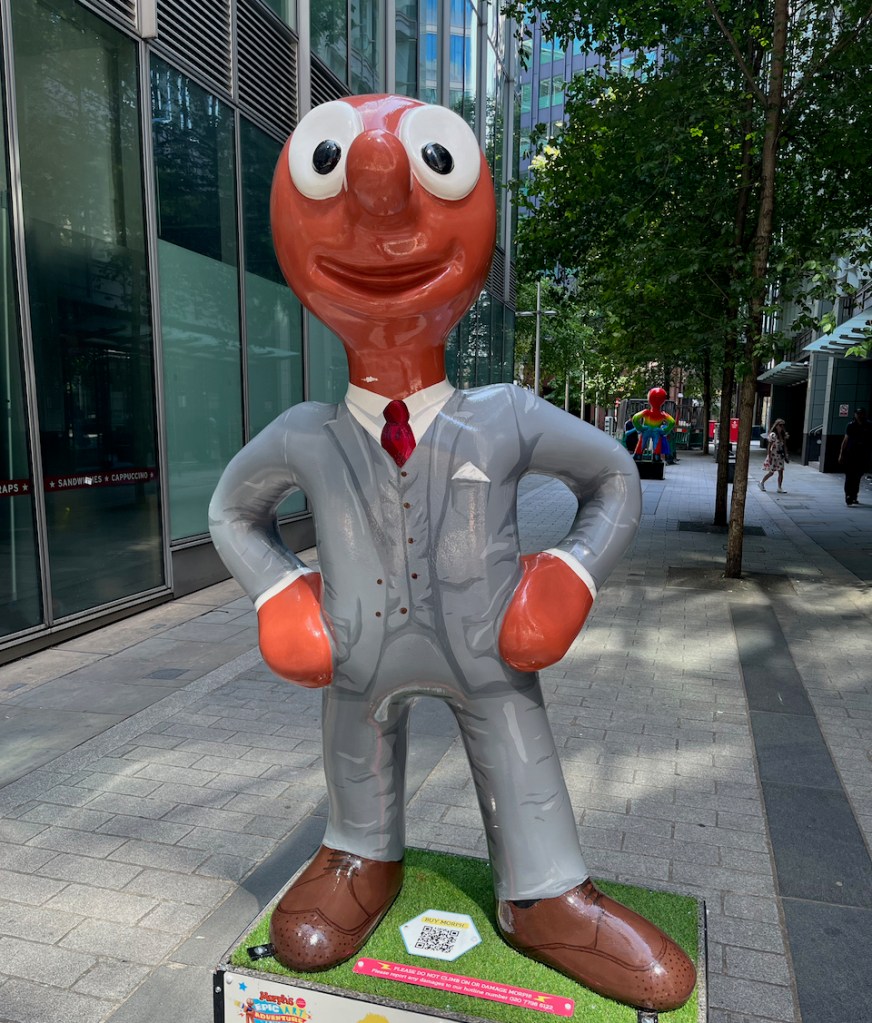 Gentle-morph, a statue of Morph dressed in a smart grey suit, over a white shirt with a red tie, and brown shoes.