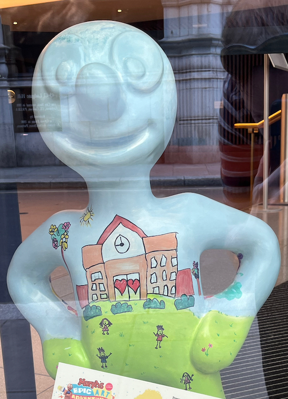 Growing Together in Learning and in Faith, a mini statue of Morph, whose light blue body features a drawing of children playing on grass outside a school building that has 2 hearts as the entrance doors.