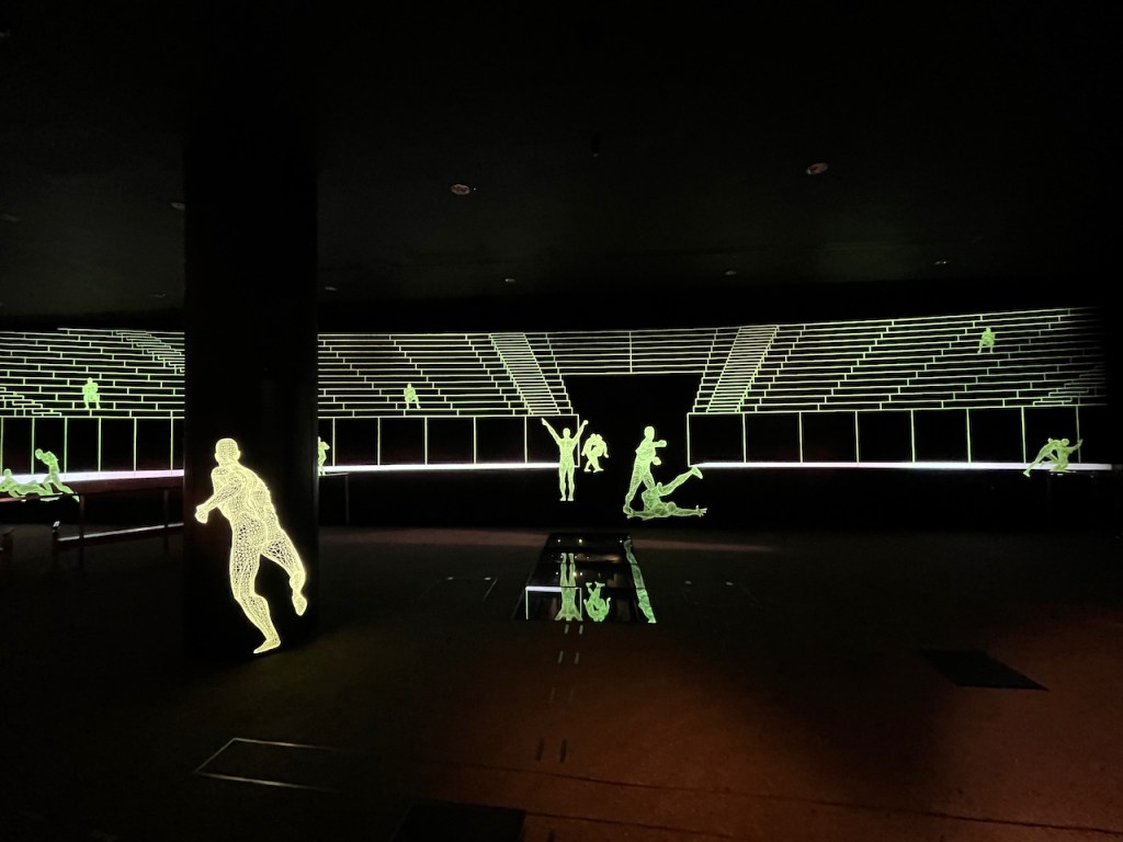 The Roman amphitheatre beneath Guildhall Art Gallery. The open plan space has green lighting across the back wall sketching out the shape of the stands where the audience would have been seated, while there are figures engaging in athletic or combative activities standing around the floor, lit in a similar way.