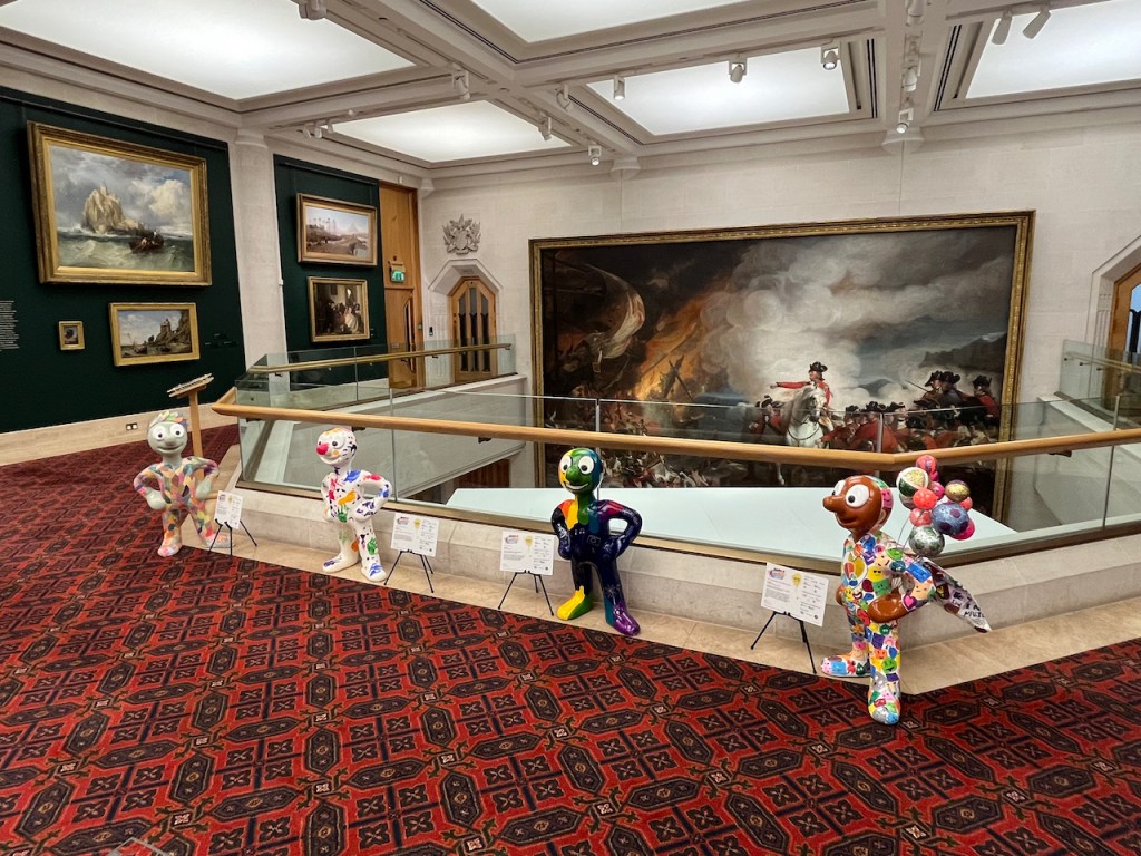 4 colourful Mini Morph statues in Guildhall Art Gallery, by a railing over which you can look at huge painting about a battle at Gibraltar.