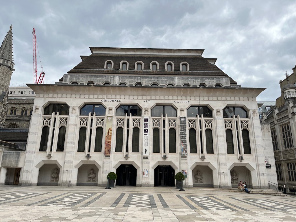 The Guildhall Art Gallery building. The upper floor is lined with 6 archways that each contain a pair of tall narrow windows, while the ground floor contains 6 open archways, 4 of which have stone on busts on the walls behind them, while the middle 2 lead to the entry and exit doors of the gallery.