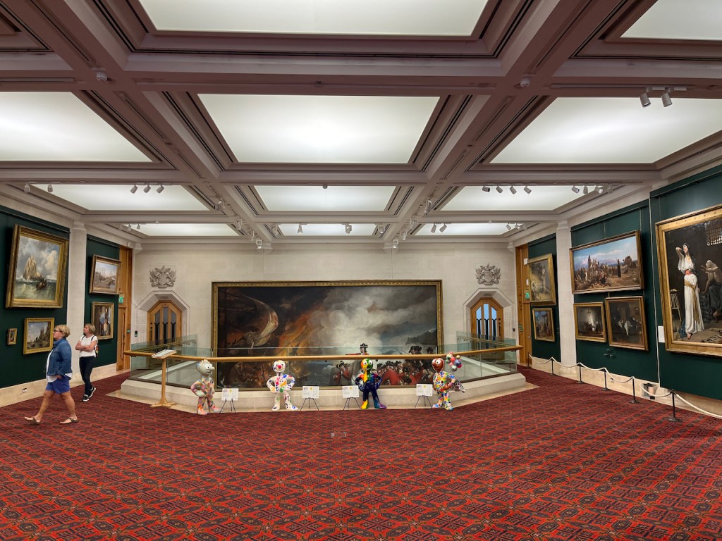 The Victorian gallery in the Guildhall Art Gallery, a large red carpeted space with paintings on the walls, and square light panels on the ceiling. At the end of the room are 4 mini statues of Morph in front of a railing, beyond which is an open space in the floor. On the wall at the back of that space is the top half of a huge painting that spans two floors of the building.