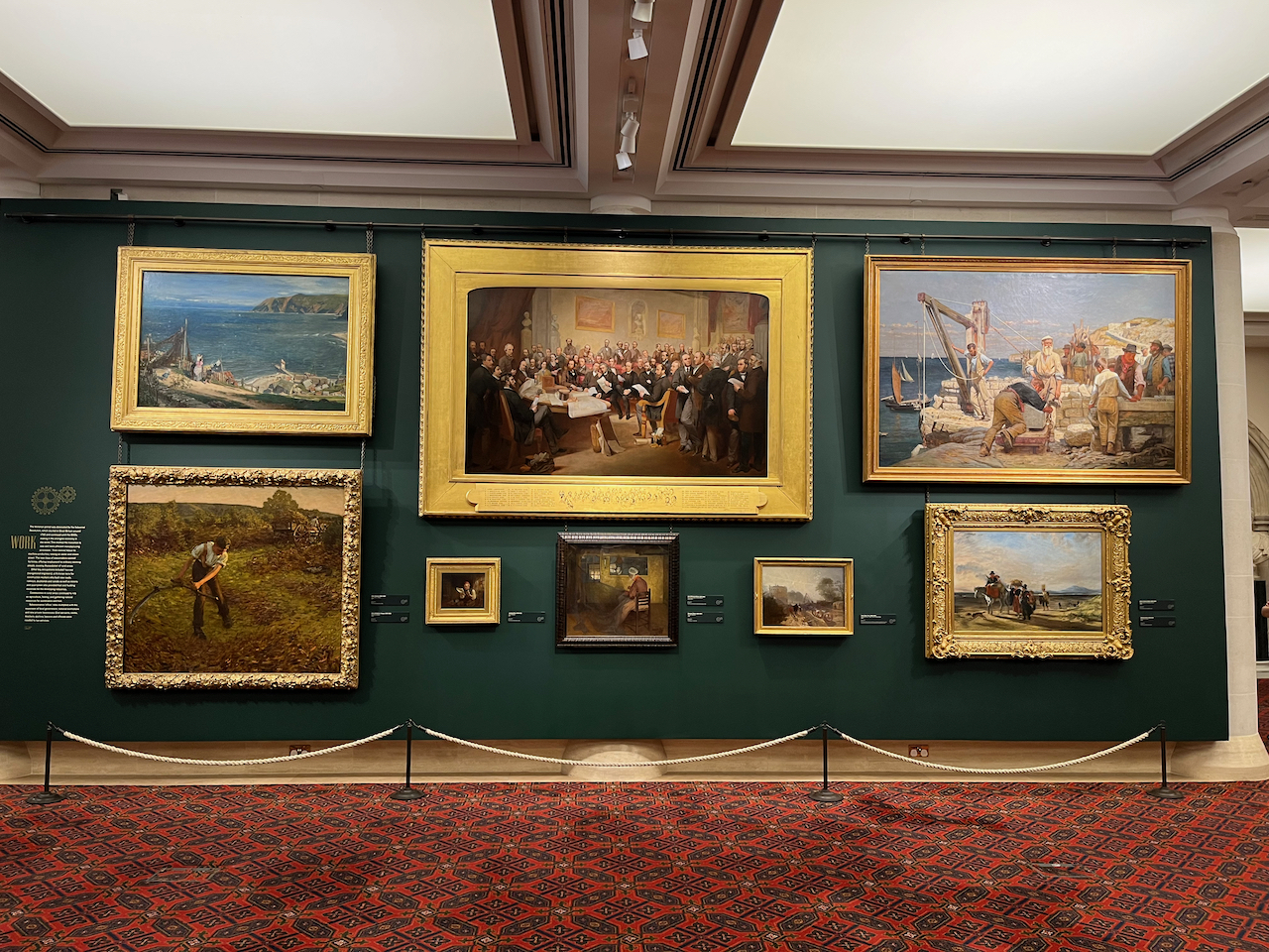 Paintings on the wall of the Victorian gallery in the Guildhall Art Gallery, including scenes by the sea and in fields, and a gathering of smartly dressed gentlemen in a large room.