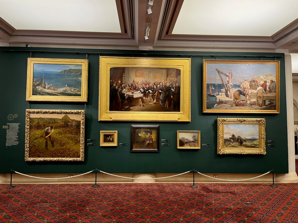 Paintings on the wall of the Victorian gallery in the Guildhall Art Gallery, including scenes by the sea and in fields, and a gathering of smartly dressed gentlemen in a large room.