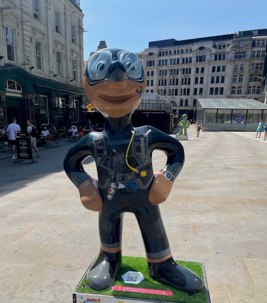 Looking After The Ocean, a statue of Morph dressed in a navy-coloured diver's wetsuit, with a breathing tube and dials on it, a watch on his wrist, and a large pair of glass goggles over his eyes.
