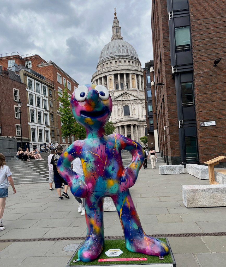 Make Your Mark, a statue of Morph covered from head to know in random splashes of colour, including pinks, blues, reds, yellows and greens, all blending into each other. St Paul's Cathedral stands tall at the end of the street in the background.