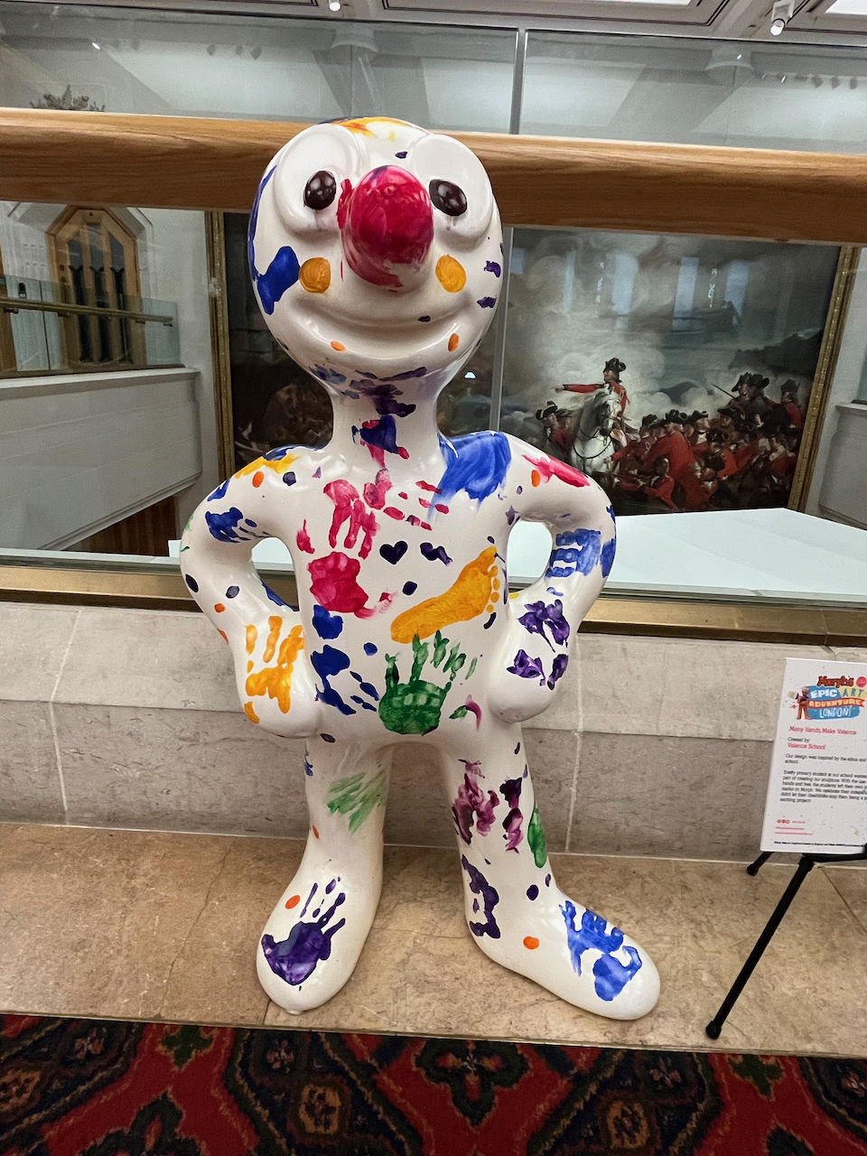 Many Hands Make Valence, a mini statue of Morph, whose white body is covered in messy colourful prints of children's hands and feet.