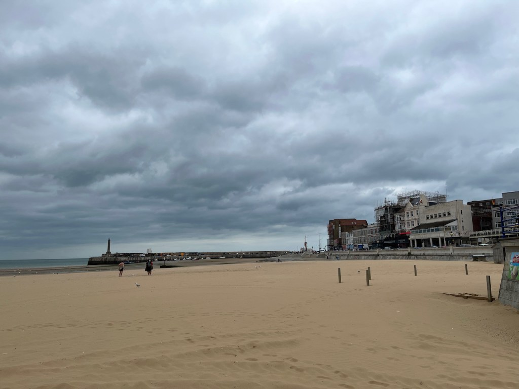 The beach at Margate. In the background, buildings line the far side of a road on the right, while on the left there's a wall stretching a short way out to sea with a small lighthouse at the end.