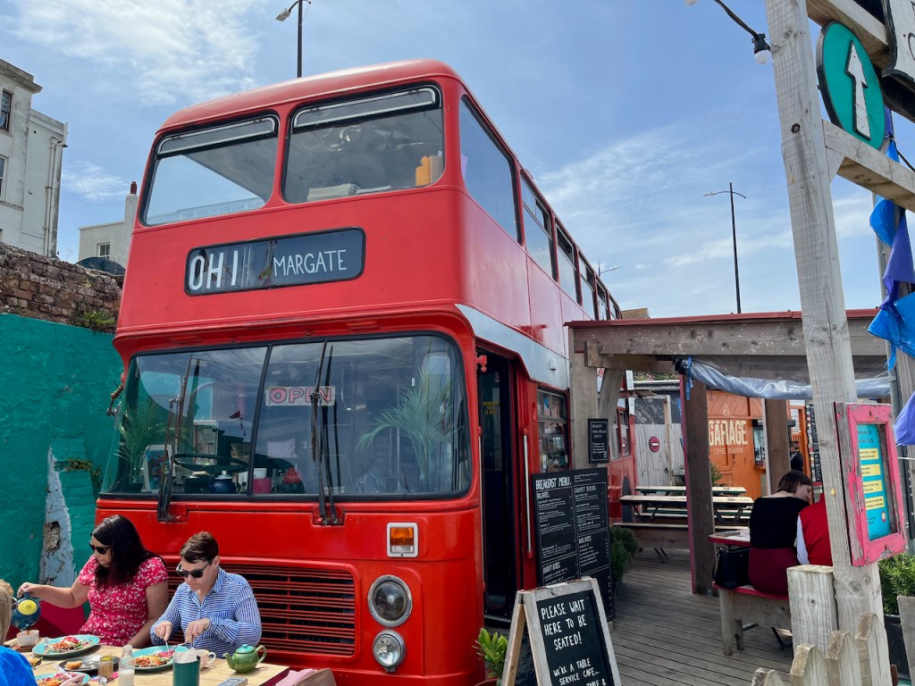 A red double-decker bus, called the Bus Café. The black destination board on the front of the bus says OH 1 Margate. A sign saying Open hangs inside the window on the bottom floor, and a few people can be seen eating at a table in the sunshine in front of the bus.