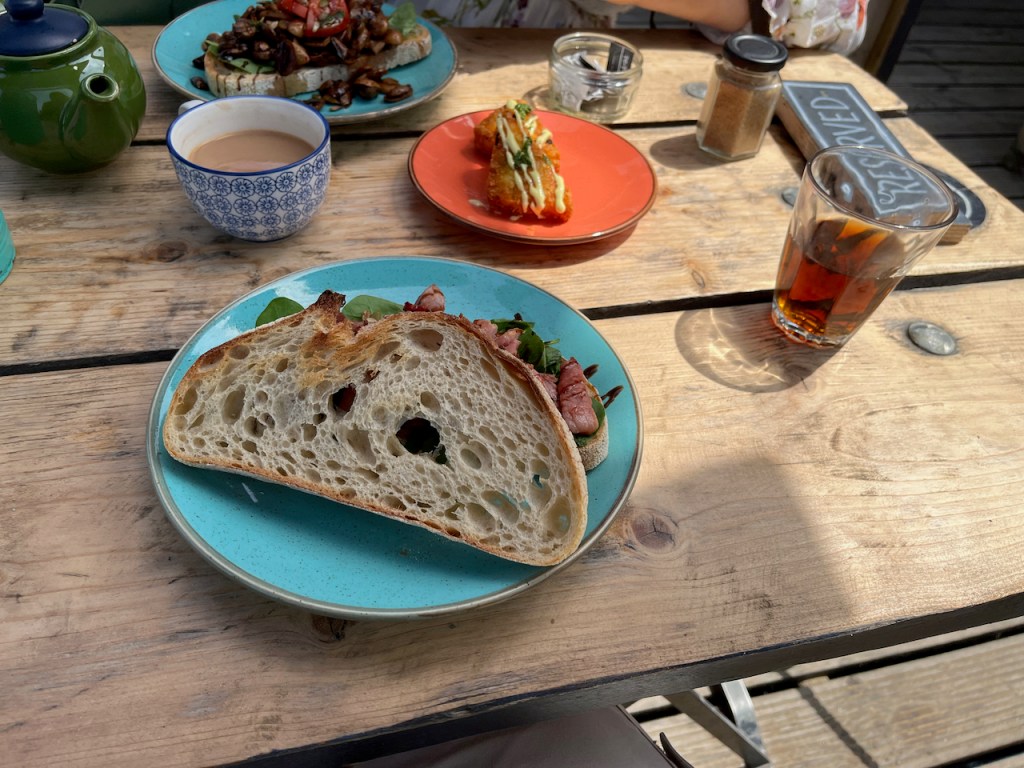 A large sourdough bacon sandwich, a side plate with a couple of has browns and a glass of cola.