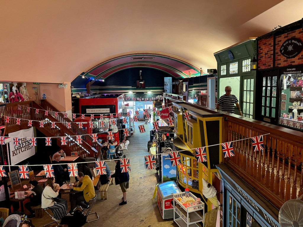 An indoor market, called The Old Kent Market, with shops and stalls selling food and other items. Strings of small Union Jack flags hang across the space at regular intervals. The photo is taken from the upper floor looking down.
