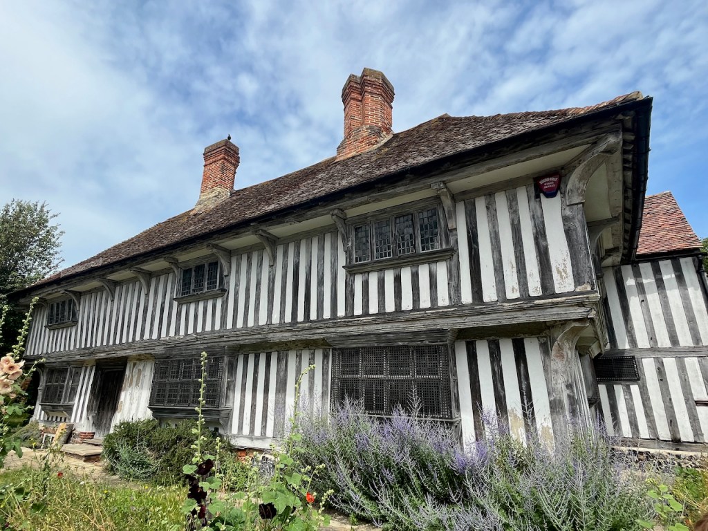 A large 2-storey Tudor House, with vertical black and white stripes all the way along the building, and 2 chimneys on the roof.