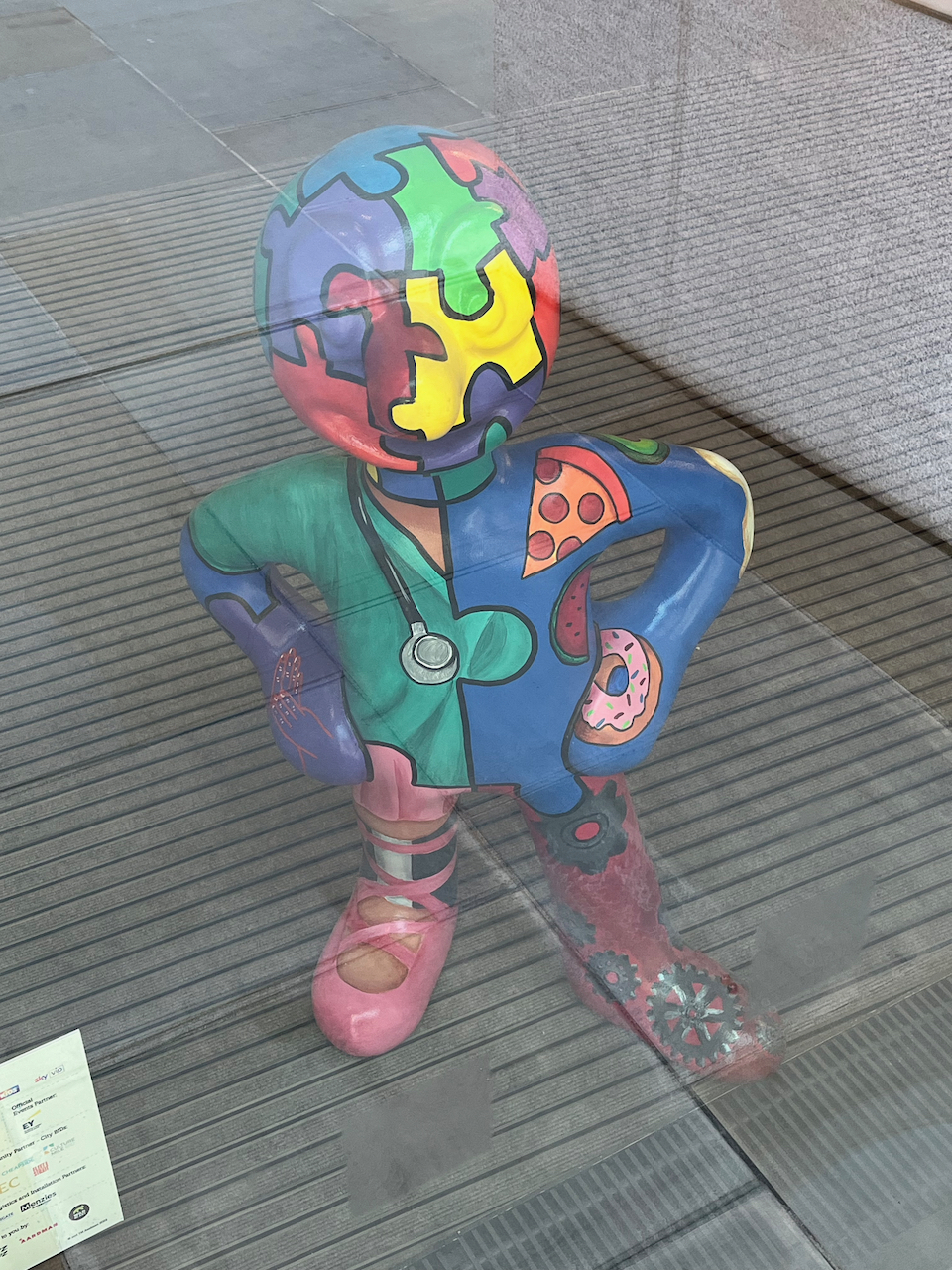 Meta-Morph-Osis, a statue of Morph whose body is made up of colourful jigsaw pieces locked together. He also has a stethoscope around his neck, a pizza slice on his left shoulder, and a ring doughnut on his left hand.