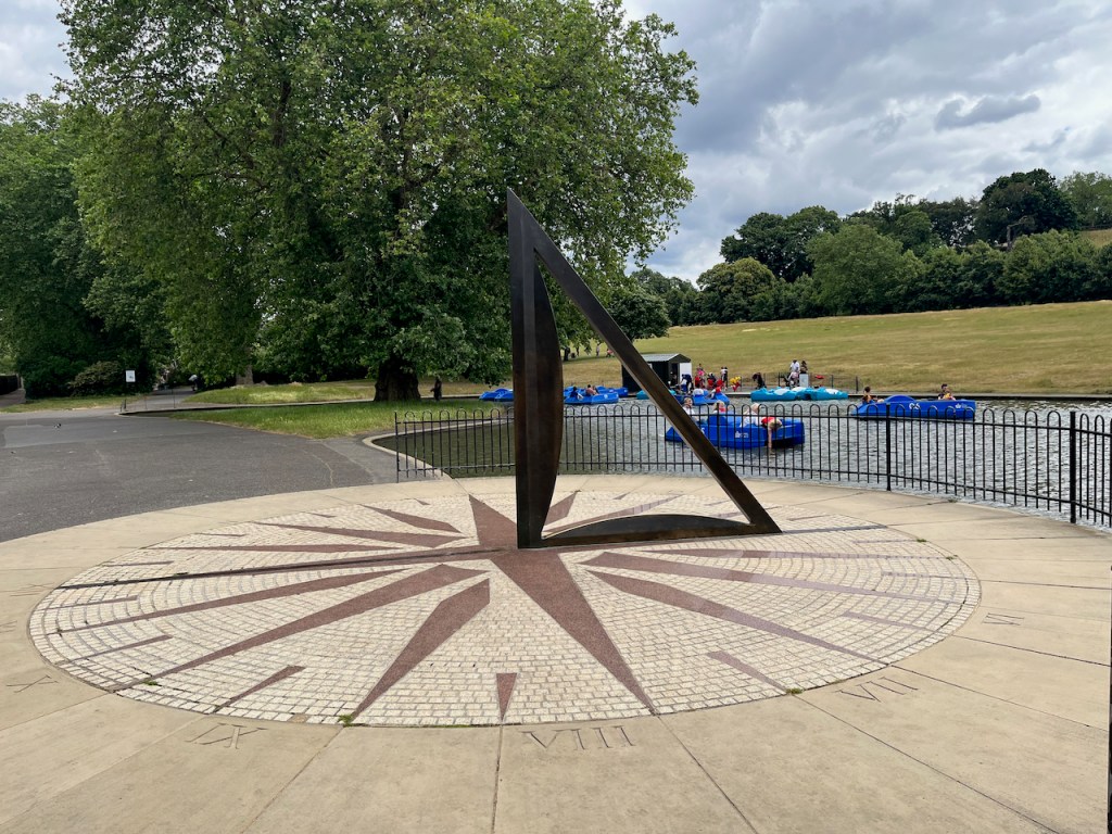 A large sundial on the ground next to a boating lake.