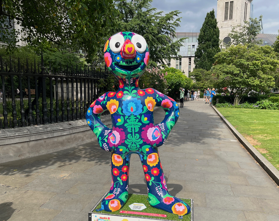 Morphlowers Please, a statue of Morph whose dark purple body is covered in bright colourful flowers and green leaves.