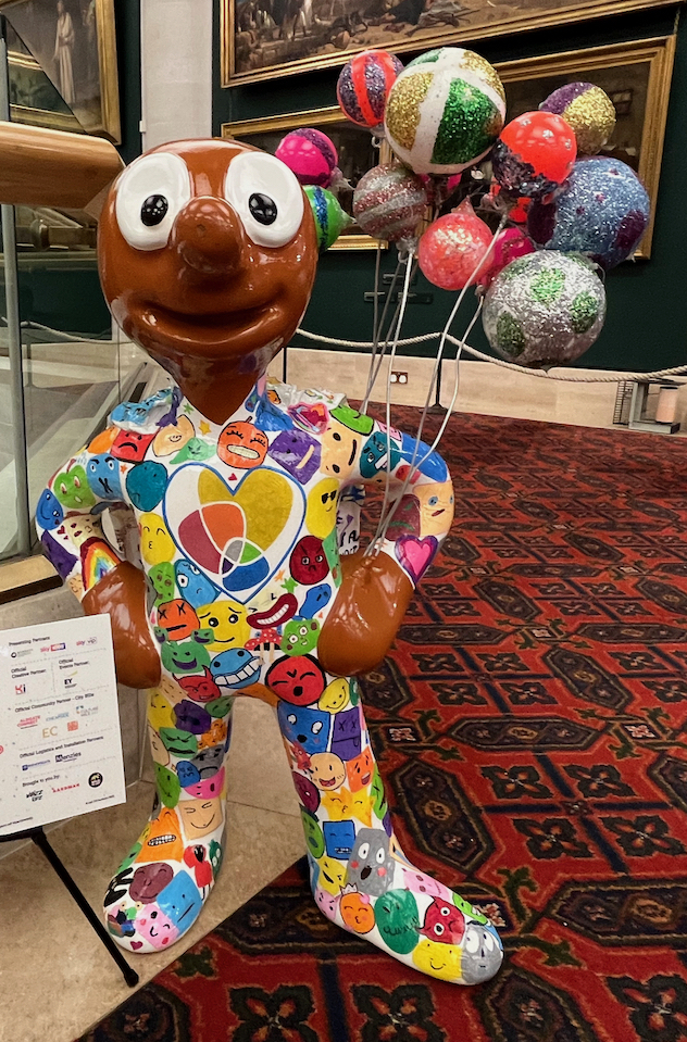 Mr Tayo Shnubbub - The Wellbeing Hero, a mini statue of Morph holding a bunch of colourful balloons, and wearing an outfit covered in lots of colourful smiling faces and hearts drawn by school children.
