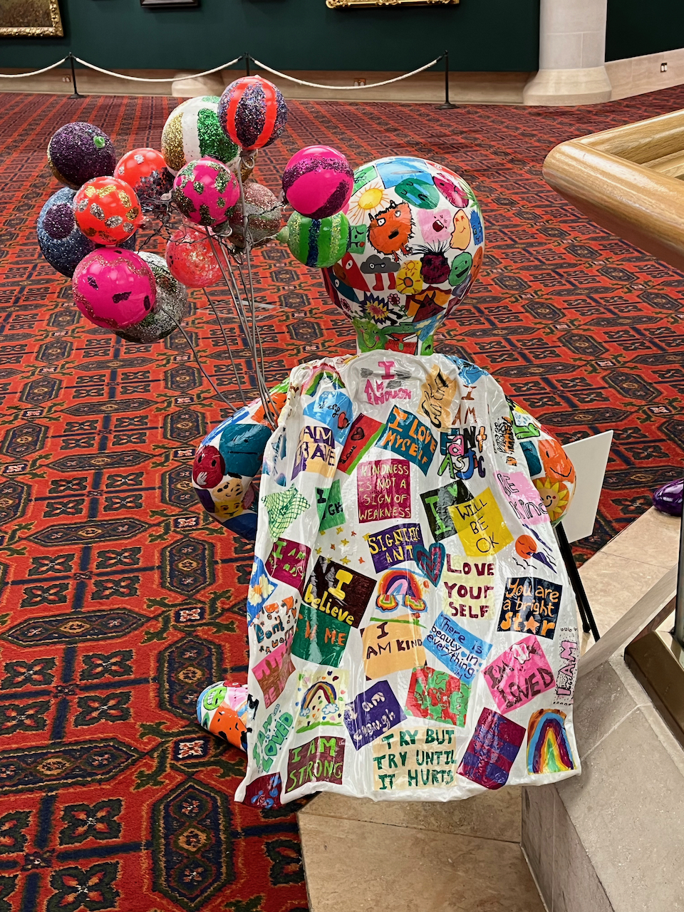 The back of Mr Tayo Shnubbub - The Wellbeing Hero, a mini statue of Morph holding a bunch of colourful balloons. The cape on his back is covered with lots of colourful messages drawn by school children, such as I am fantastic, I am enough, kindness is not a sign of weakness, will be ok, love yourself, you are a bright star, I am kind, there is beauty in everything, I am strong, try but try until it hurts, and I'm loved.