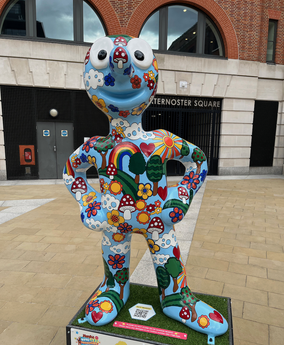 Peace, Love and Morph, a statue of Morph whose light blue body is covered in colourful rainbows, mushrooms, flowers, trees and clouds.