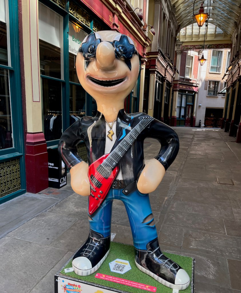 Rock Star, a statue of Morph dressed as a rock musician, with black star-shaped sunglasses, a small gold necklace shaped like a lightning bolt, a black leather jacket over a white t-shirt, blue jeans, black trainers with white toecaps, and a red guitar held diagonally across his body.