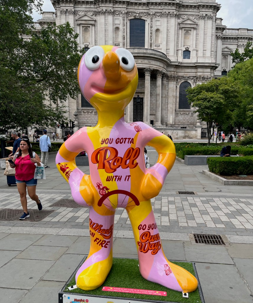 Roll With It, a statue of Morph painted in alternating yellow and pink colours. Text on his chest reads You gotta roll with it, above a small image of a wheelchair user riding on a curve over Morph's lower chest. Text on each of Morph's legs reads Wheels on fire and Go your own way.