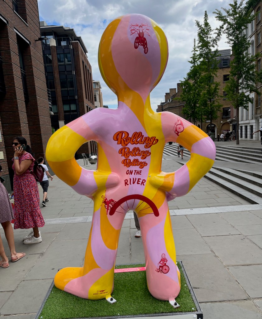The back of Roll With It, a statue of Morph painted in alternating yellow and pink colours. Text on his back reads Rolling, Rolling, Rolling on the river, above a small image of a wheelchair user riding on a curve over Morph's bottom. A small image of a wheelchair user also appears just above the heel of Morph's right leg.