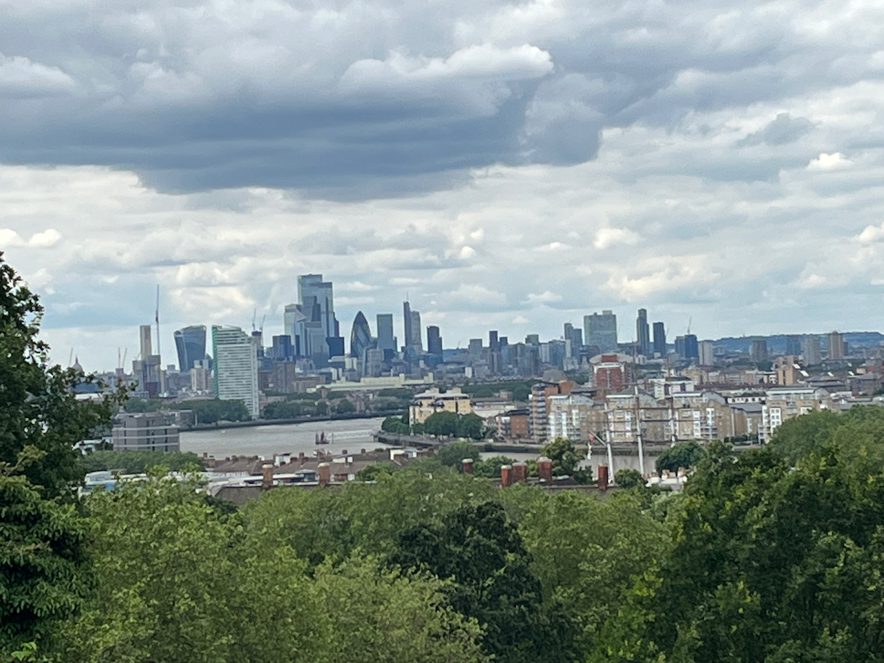 View looking across trees towards the River Thames as it curves to the right. The tall buildings in the London skyline behind it include the so-called Walkie Talkie and Gherkin buildings.