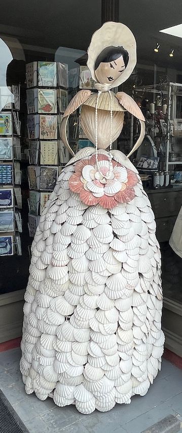 Statue of a lady outside the entrance to the Shell Grotto. Three quarters of her frame is a huge skirt made of layers of white shells, above which is a small torso with thin arms, and a head wearing a bonnet.