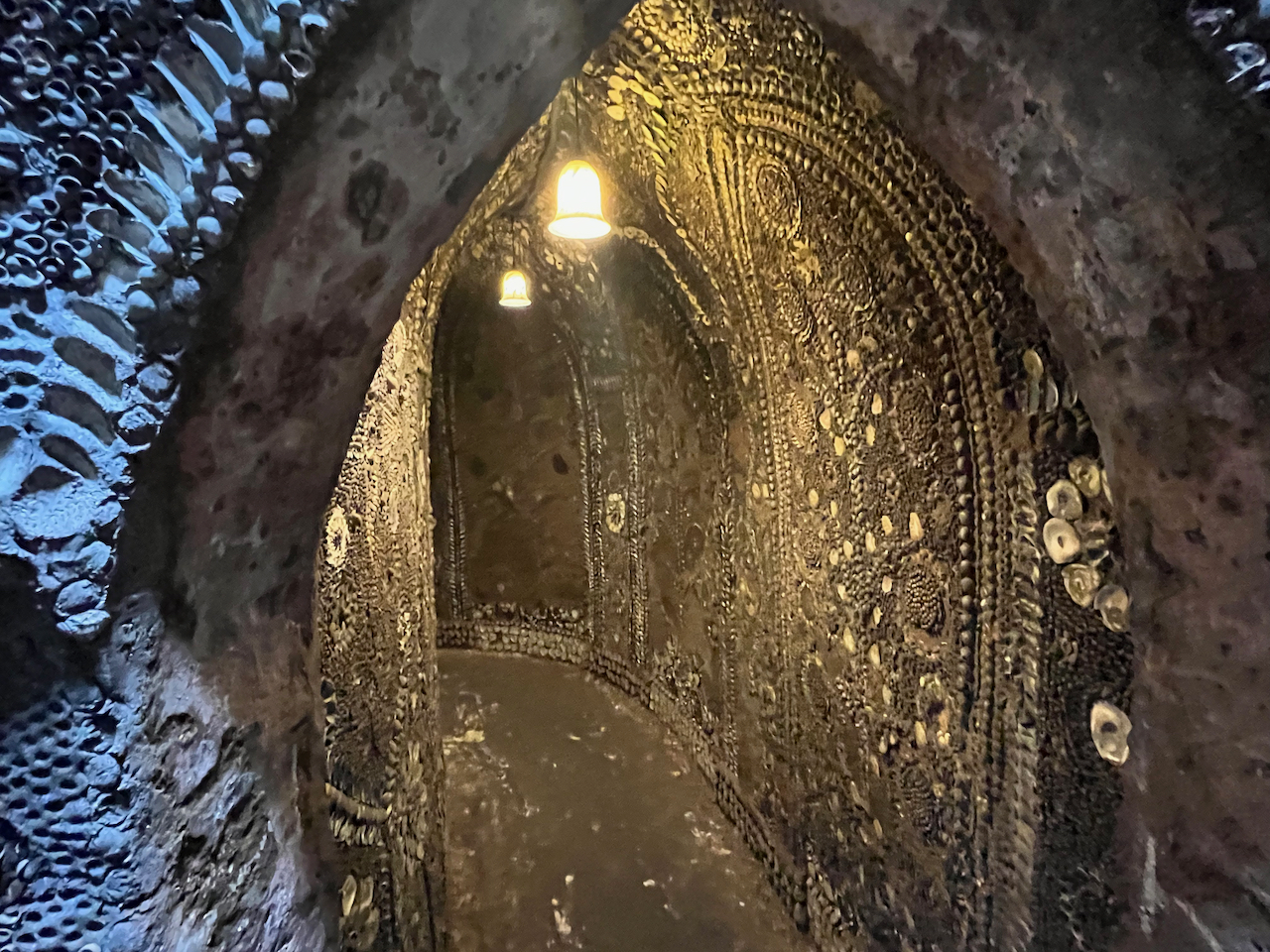 A narrow corridor with intricate patterns made of shells on the walls, and a couple of small lights hanging from the ceiling.
