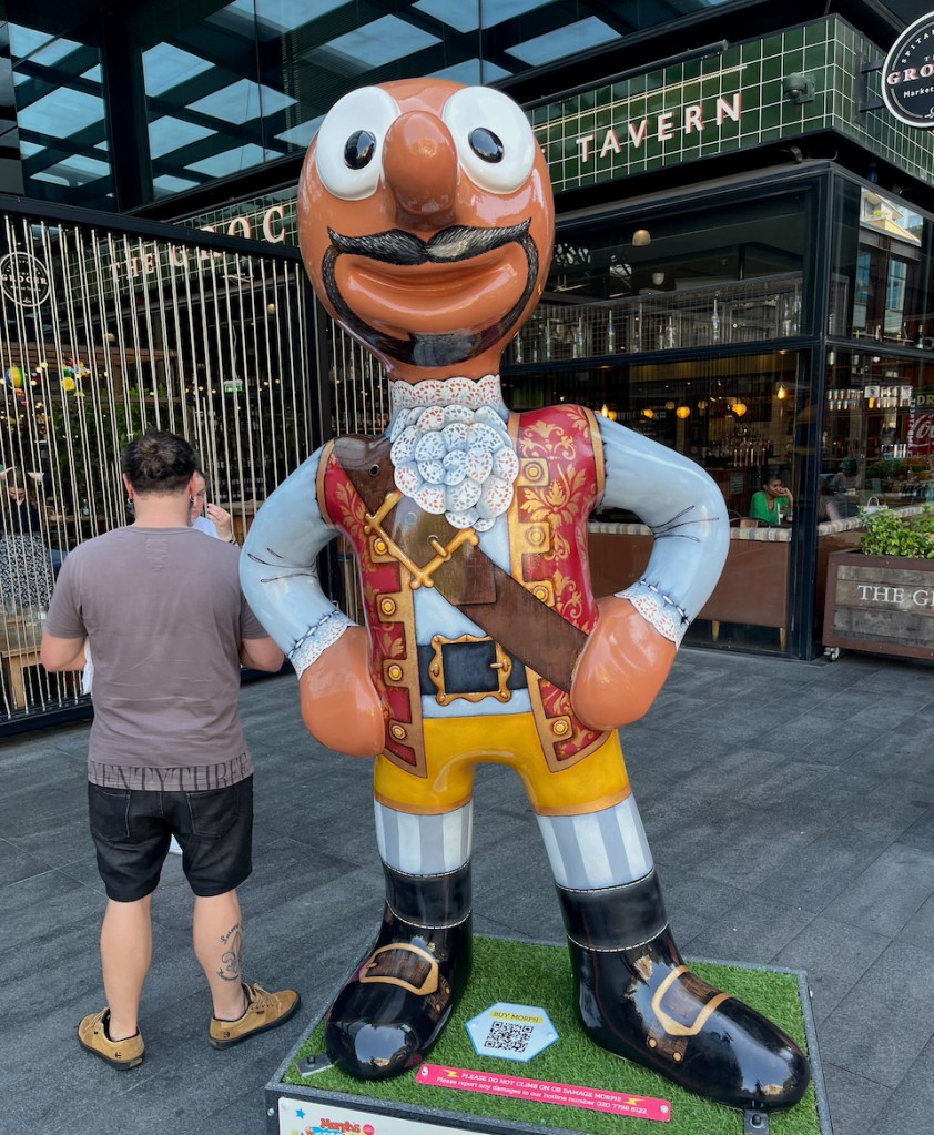 Swashbuckler, a statue of Morph dressed as a pirate, including a sleeveless red jacket with ornate gold detailing over a white shirt, a black belt around his waist under the jacket, and a thick brown belt stretched diagonally from one shoulder down across his chest. He's also wearing yellow shorts, grey and white striped socks and black boots, and has a black beard and moustache.