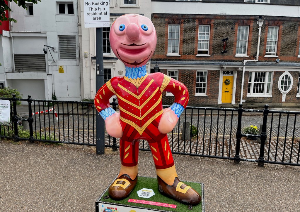 The Bard, a statue of Morph wearing a red jacket and trousers with gold stripes and gold trim, a blue ruff around his neck, brown shoes with gold buckles, and on his face he has a very thin black beard and moustache.