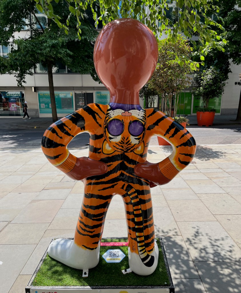 The back of the Tiger Morph statue, which shows the back of Morph's orange tiger onesie with black stripes. An upside down tiger's head with large holes for the eyes hangs down from Morph's neck, as if it's a hood he can pull over his head to show the face of a tiger.