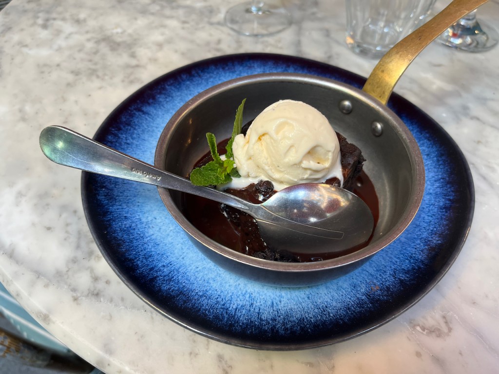 Dessert at Bill's restaurant in Covent Garden, consisting of a triple chocolate brownie with a blob of vanilla ice cream on top. It's served in a dark coloured bowl with a long, slightly curving handle on one side, making it look like a bit like a very small and very shallow frying pan.
