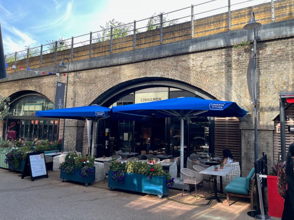 Cinnamon Kitchen restaurant, underneath an archway of the Grosvenor Railway Bridge. There are some seats and tables outside the front of the restaurant, underneath a flat blue canopy.