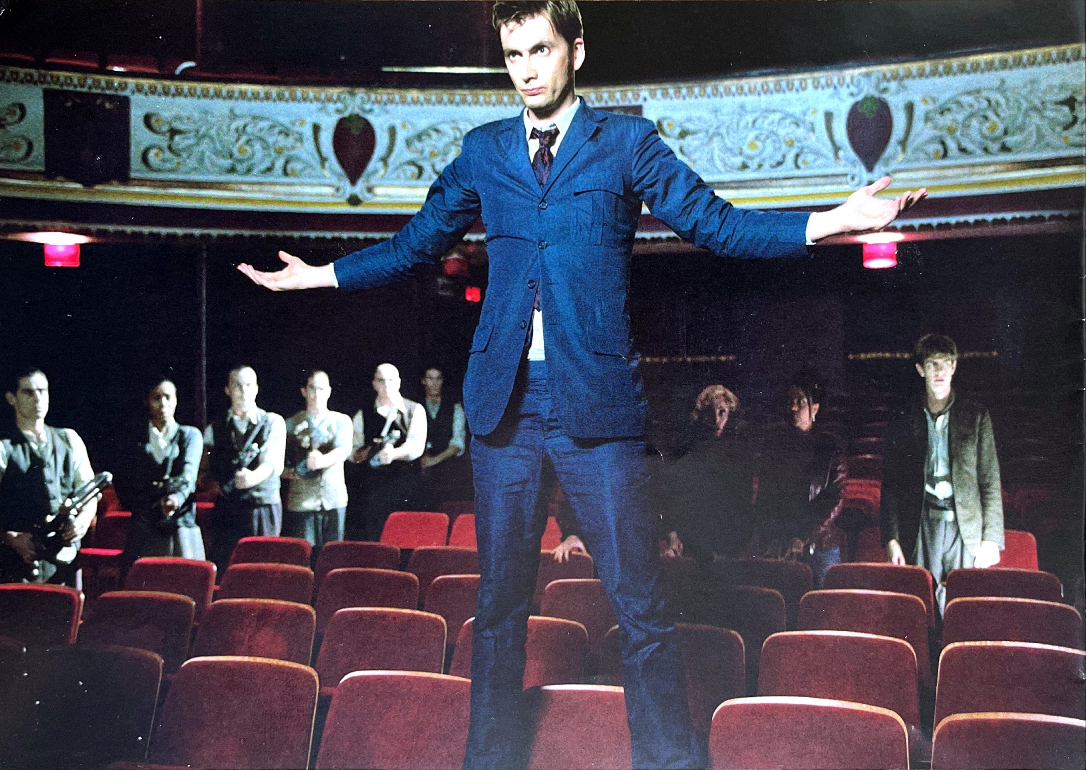 David Tennant's Doctor in a blue suit, standing in the middle of a row of red seats in a theatre auditorium, his arms stretched out to his sides. In the aisle to the left, a line of people hold Dalek guns pointed towards him. A few rows behind the Doctor, Martha Jones stands anxiously with Tallulah and Frank.