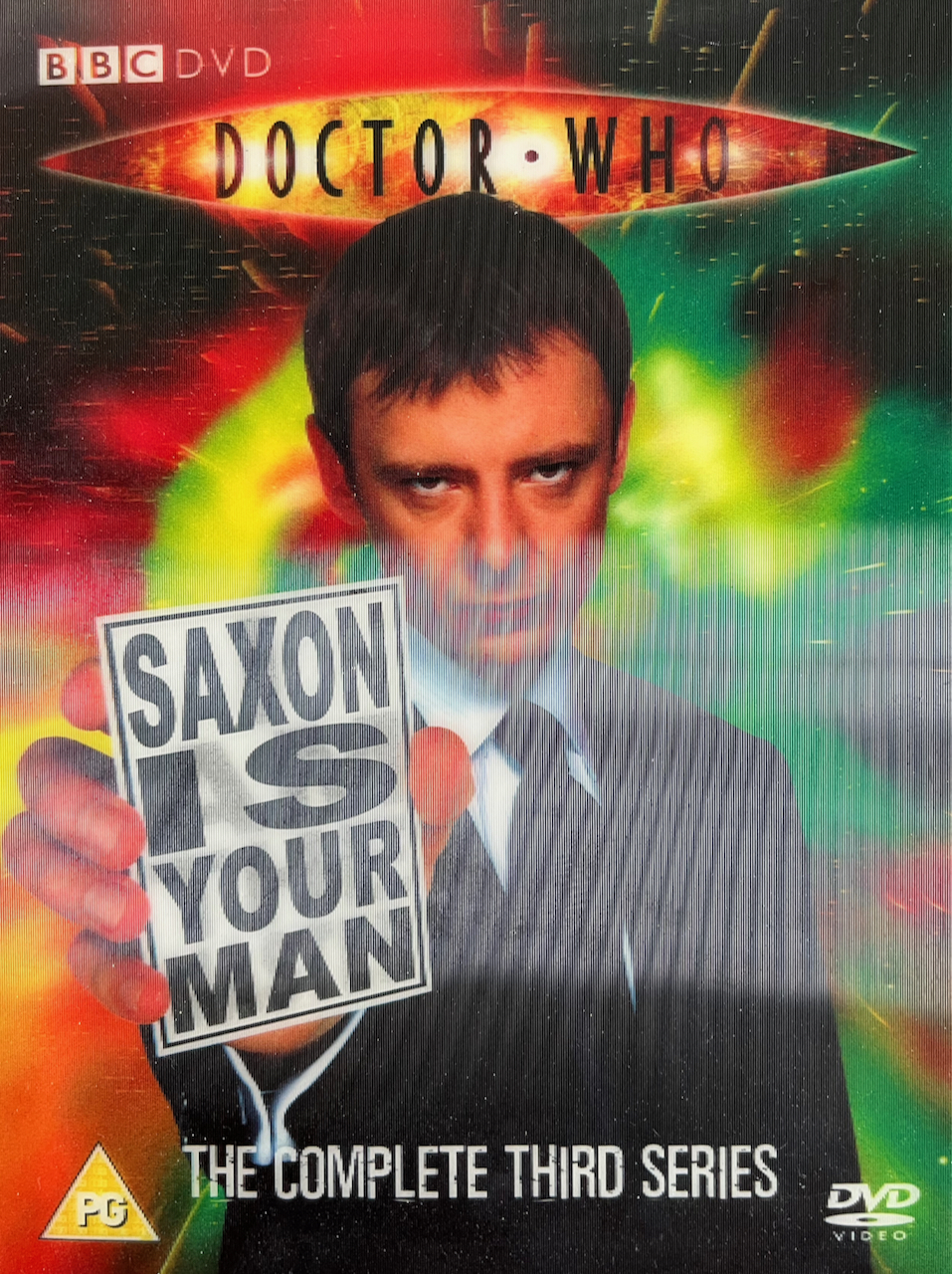 Lenticular cover art from the old Doctor Who Series 3 DVD set, featuring John Simm as The Master holding up a piece of white card with large black lettering that reads Saxon Is Your Man. In fainter black lettering behind it are the words I Am The Master, which can be seen when tilting the case to a different angle.